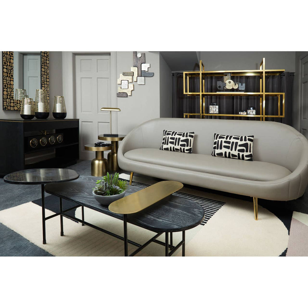 Brown leather effect 3 seater sofa with a curved backrest, placed in a modern living room setting. The sofa features decorative cushions with a geometric pattern. A sleek black coffee table with a wooden accent sits in front of the sofa, surrounded by a light-colored rug. The background includes a stylish shelving unit and wall decor, creating a contemporary atmosphere