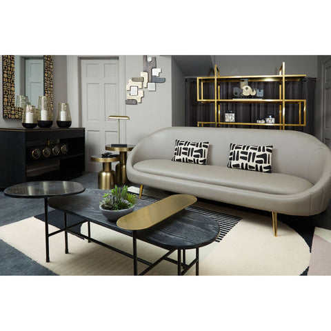 Brown leather effect 3 seater sofa with a curved backrest, placed in a modern living room setting. The sofa features decorative cushions with a geometric pattern. A sleek black coffee table with a wooden accent sits in front of the sofa, surrounded by a light-colored rug. The background includes a stylish shelving unit and wall decor, creating a contemporary atmosphere