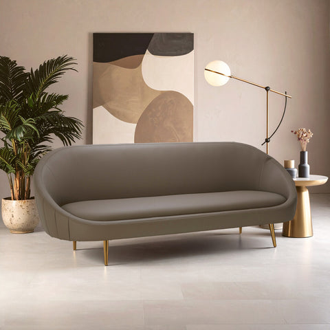 Brown leather effect 3 seater sofa with a curved design, featuring a smooth surface and gold metal legs. The sofa is positioned in a modern living space with a large abstract wall art piece, a stylish floor lamp, and a decorative plant in the background. The floor is light-colored, enhancing the contemporary aesthetic of the room