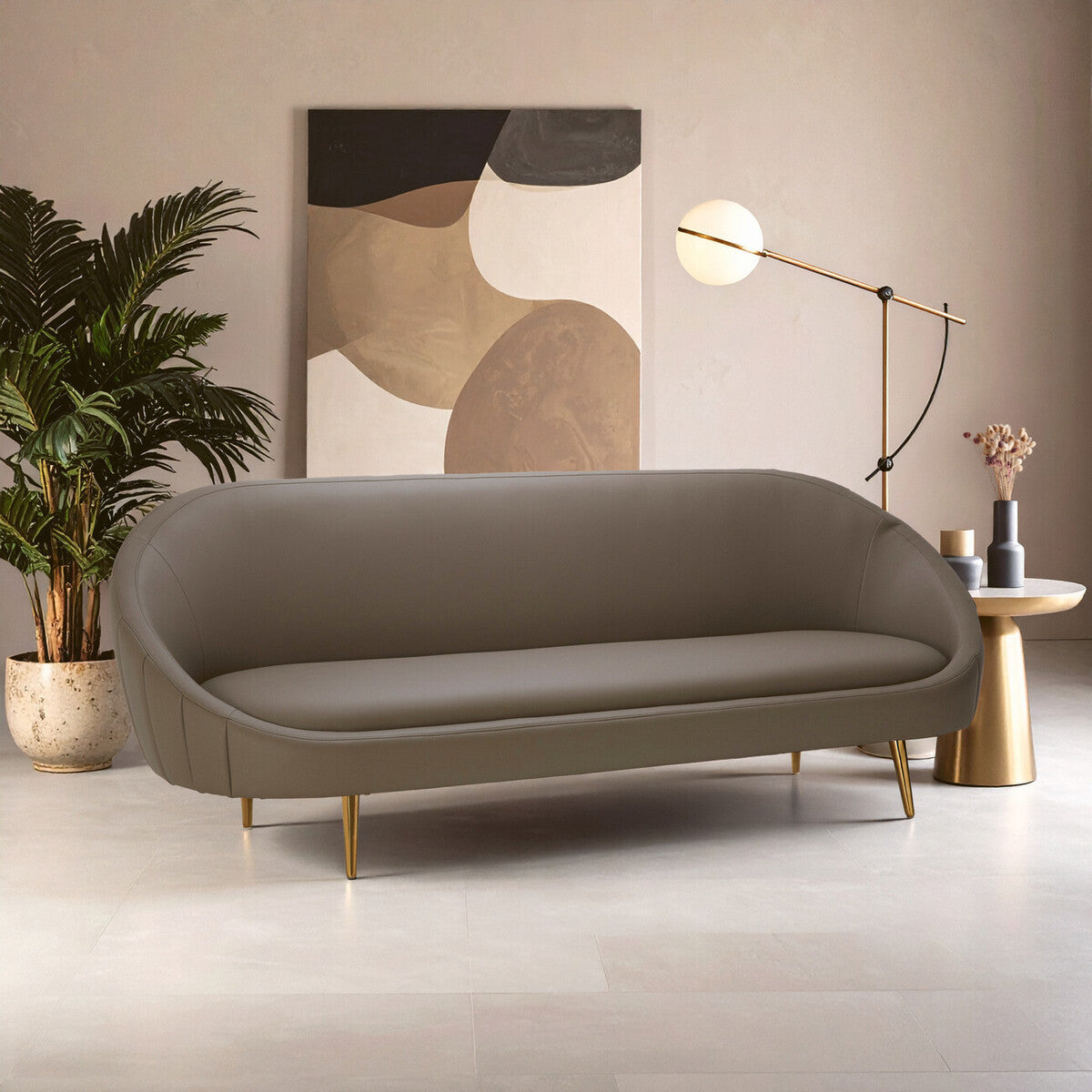 Brown leather effect 3 seater sofa with a curved design, featuring a smooth surface and gold metal legs. The sofa is positioned in a modern living space with a large abstract wall art piece, a stylish floor lamp, and a decorative plant in the background. The floor is light-colored, enhancing the contemporary aesthetic of the room