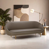 Brown leather effect 3 seater sofa with a curved design, featuring a smooth surface and gold metal legs. The sofa is positioned in a modern living space with a large abstract wall art piece, a stylish floor lamp, and a decorative plant in the background. The floor is light-colored, enhancing the contemporary aesthetic of the room