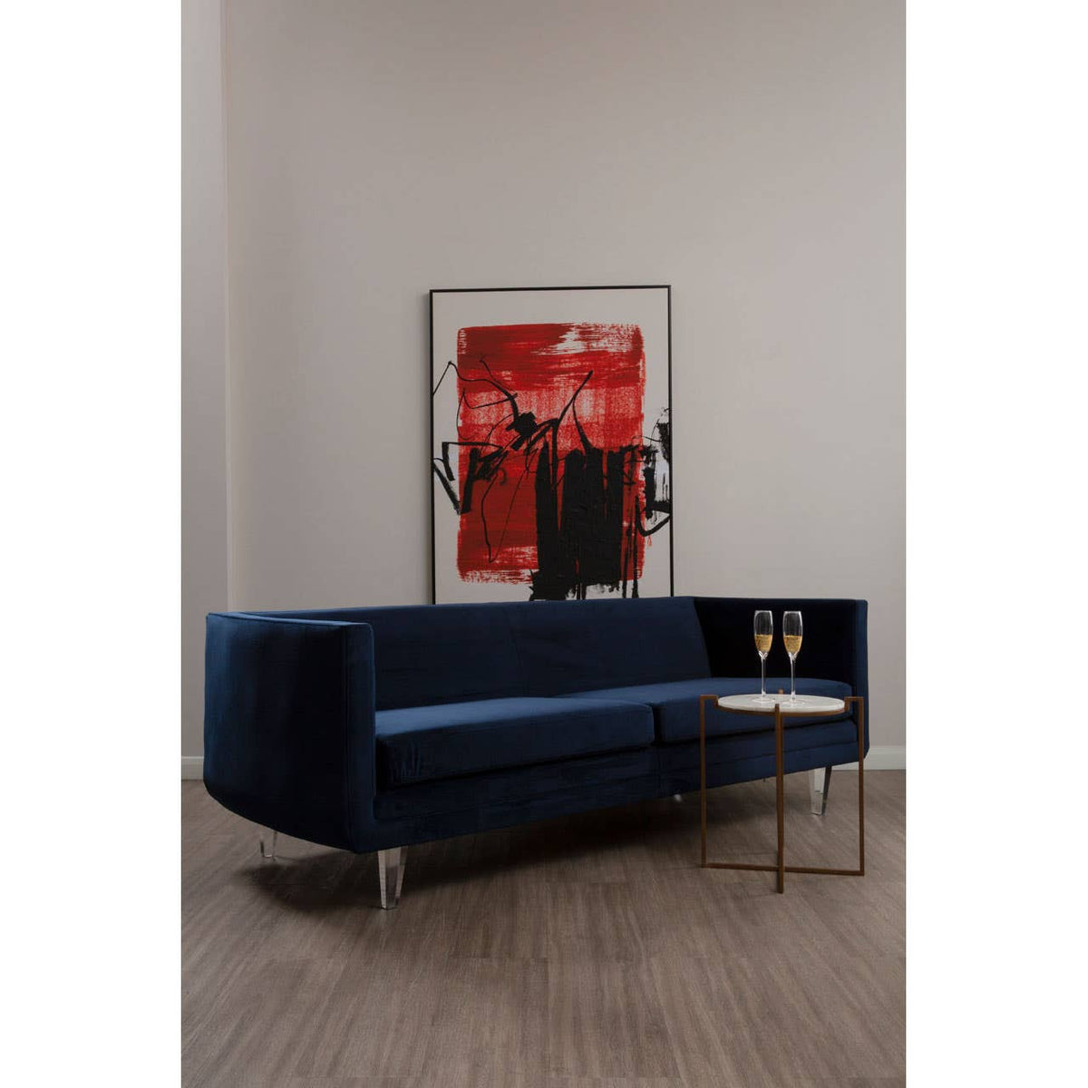 Navy velvet three-seater sofa with a sleek design, featuring a low backrest and a side table integrated into the armrest, set against a neutral wall with a bold red and black artwork in the background. The sofa is supported by slender gold legs, and the floor is made of light wood