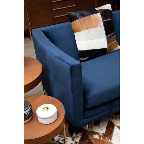 Navy velvet three-seater sofa with clean lines and a modern design, featuring a plush seat and armrests. A decorative pillow with geometric patterns is placed on the sofa. In the foreground, a round wooden side table holds a white decorative box and a small black item, while a textured rug is visible beneath the furniture