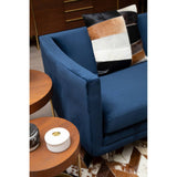 Navy velvet three-seater sofa with clean lines and a modern design, featuring a plush seat and armrests. A decorative pillow with geometric patterns is placed on the sofa. In the foreground, a round wooden side table holds a white decorative box and a small black item, while a textured rug is visible beneath the furniture
