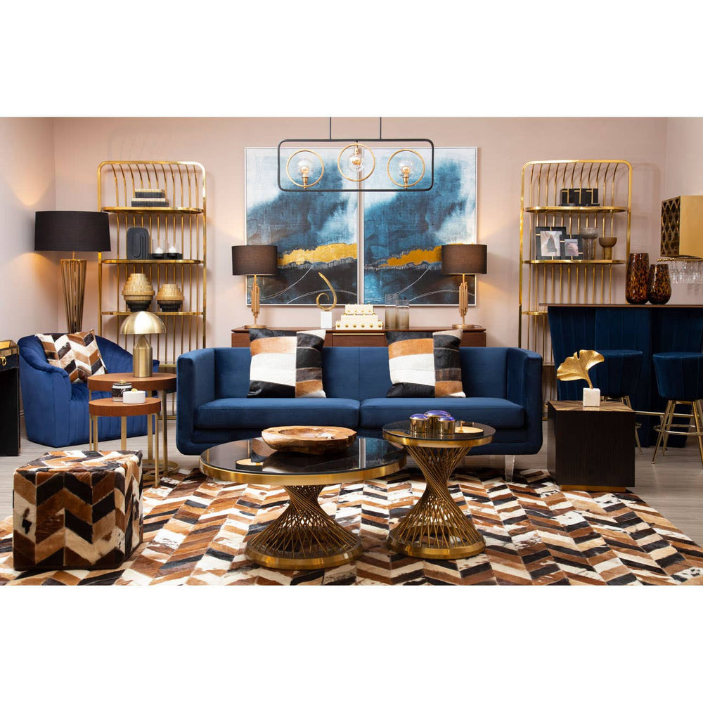 Navy velvet three-seater sofa with plush cushions, set against a stylish living room backdrop featuring light pink walls, wooden shelves, decorative items, and a patterned area rug. The sofa is flanked by two blue armchairs and complemented by a coffee table