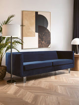 Navy velvet three-seater sofa with a sleek design, featuring a low back and open side, positioned in a modern living space with a wooden floor. A tall floor lamp and a round table lamp are visible nearby, along with an abstract wall art piece in the background. A green plant adds a touch of nature to the setting
