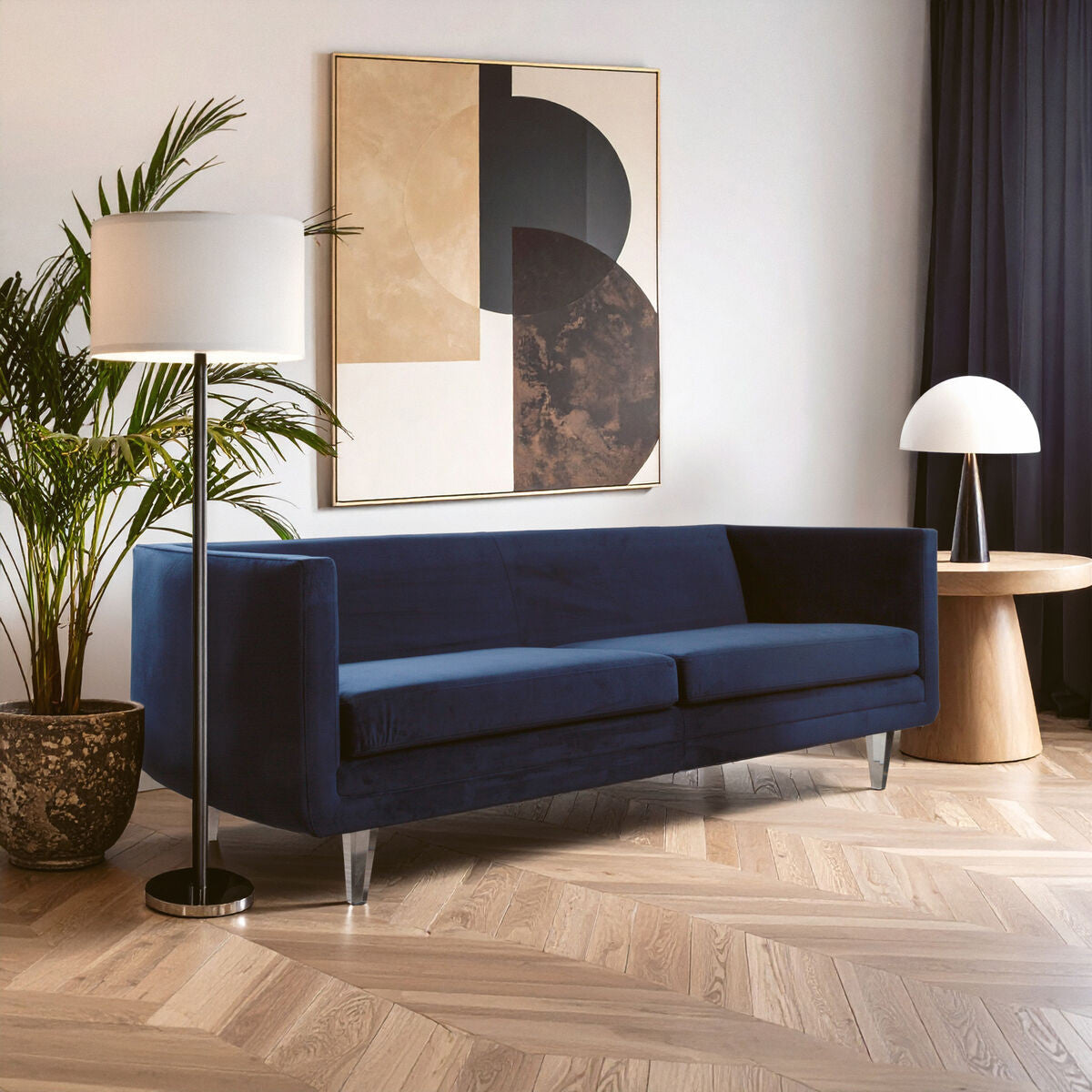 Navy velvet three-seater sofa with a sleek design, featuring a low back and armrests, positioned in a modern living room setting. The sofa is complemented by a floor lamp and a round side table, with a large abstract artwork on the wall and a potted plant nearby. The flooring is light wood, enhancing the contemporary aesthetic