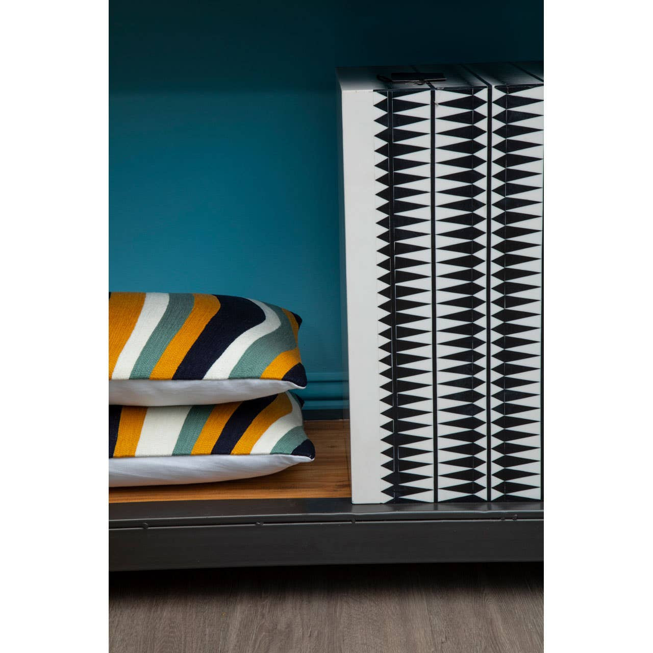 A close-up view of a shelf displaying two neatly stacked pillows with bold, striped patterns in orange, blue, and white, alongside a stack of silver, circular coasters. The background features a teal wall and a wooden shelf surface