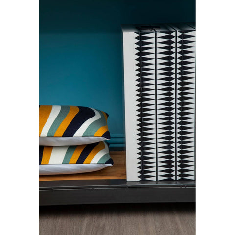 A close-up view of a shelf displaying two neatly stacked pillows with bold, striped patterns in orange, blue, and white, alongside a stack of silver, circular coasters. The background features a teal wall and a wooden shelf surface