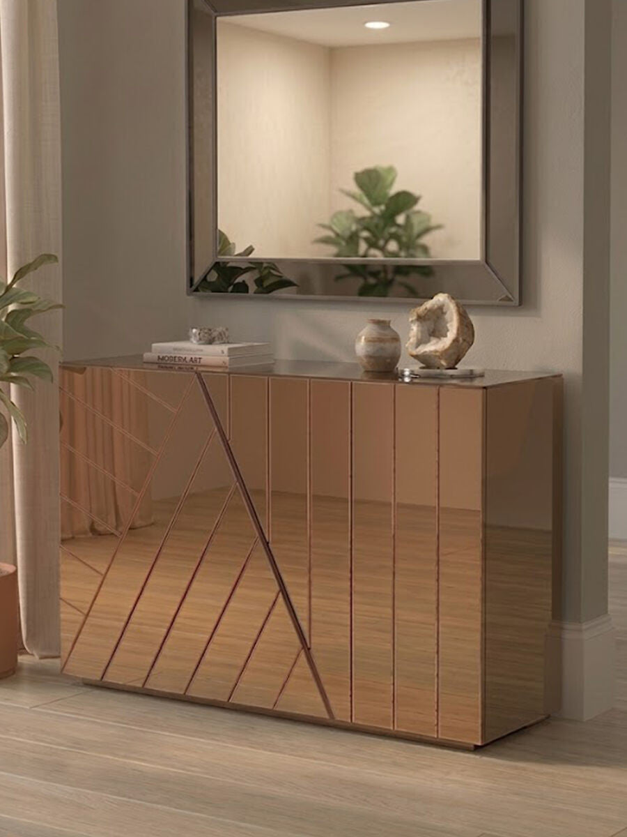 A stylish console table with a tinted mirror front and a geometric design, featuring a warm wood finish. The table is positioned against a neutral wall, with a simple decorative item on top and a large mirror reflecting the room's soft lighting. A potted plant adds a touch of greenery to the setting