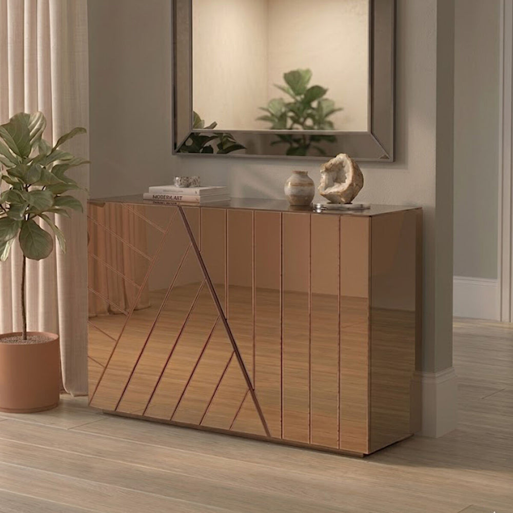 A modern console table with a tinted mirror finish, featuring a geometric design on the front. The table is positioned against a light-colored wall, with a decorative mirror above it. A potted plant is placed beside the table on the floor, enhancing the contemporary aesthetic of the room. The flooring is light wood, adding warmth to the space