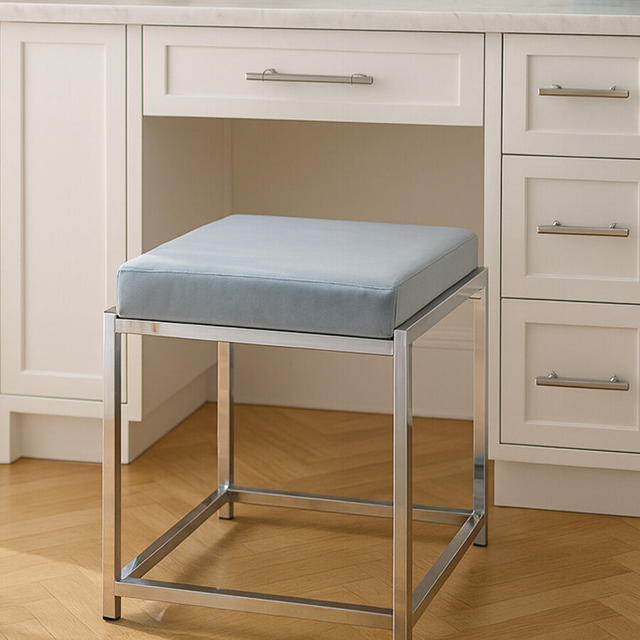 A powder blue velvet stool with a square cushion sits on a silver metal frame, positioned in front of a white desk with drawers. The floor is made of light wood, and there are decorative items on the desk, including a round vase and a small circular object