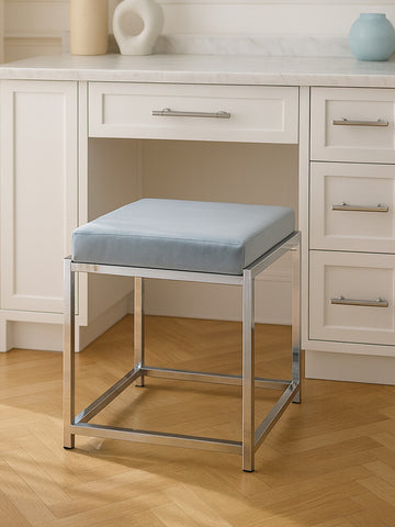 A powder blue velvet stool with a square cushion sits on a silver metal frame, positioned in front of a white desk with drawers. The floor is made of light wood, and there are decorative items on the desk, including a round vase and a small circular object