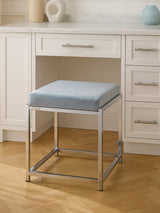 A powder blue velvet stool with a square cushion sits on a silver metal frame, positioned in front of a white desk with drawers. The floor is made of light wood, and there are decorative items on the desk, including a round vase and a small circular object