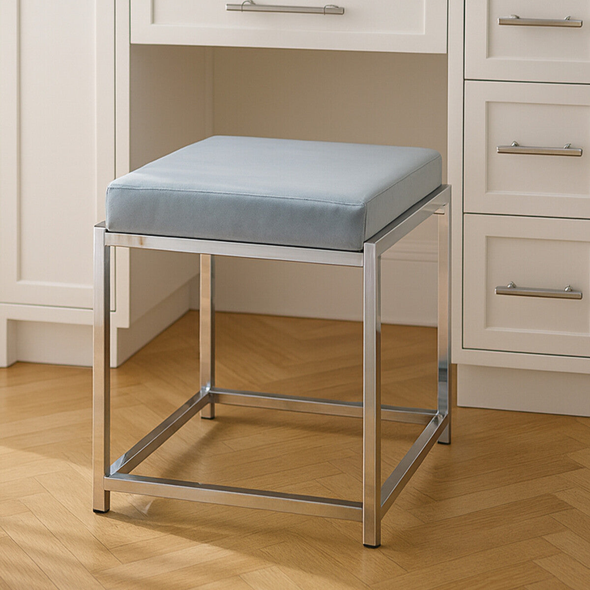 A powder blue velvet stool with a square cushion sits atop a sleek silver metal frame, featuring clean lines and a modern design. The stool is positioned on a wooden floor, with white cabinetry visible in the background