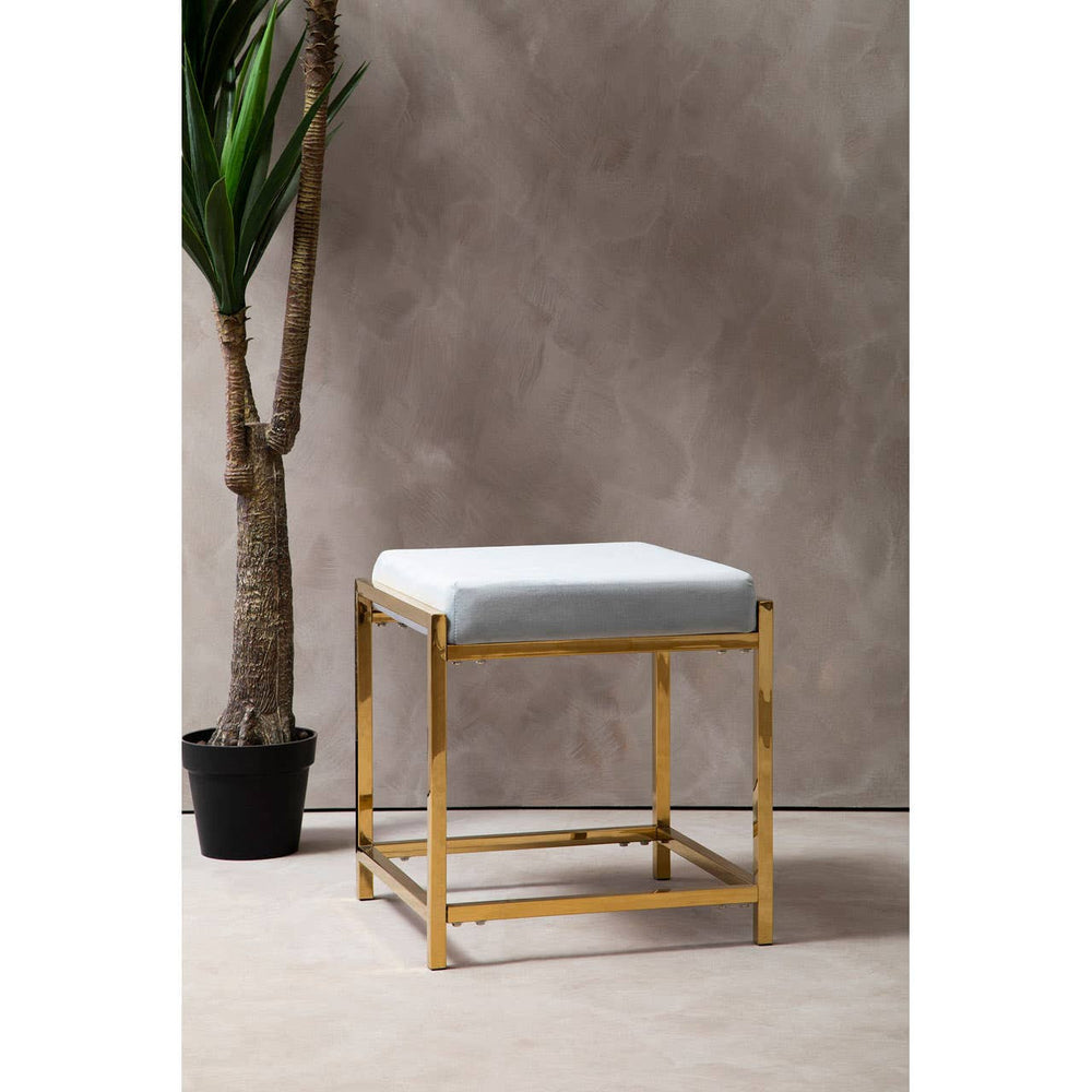 A powder blue velvet stool with a square top and a sleek gold metal frame, positioned next to a potted plant against a textured gray background