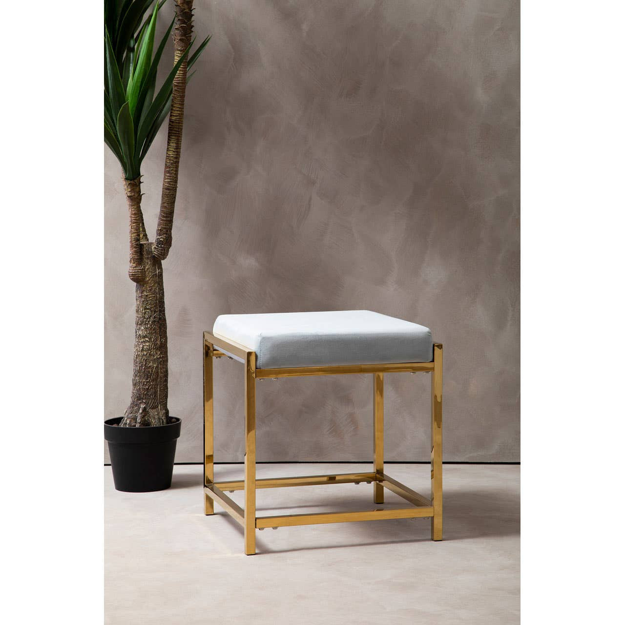 A powder blue velvet stool with a square top and a sleek gold metal frame, positioned next to a potted plant against a textured gray background