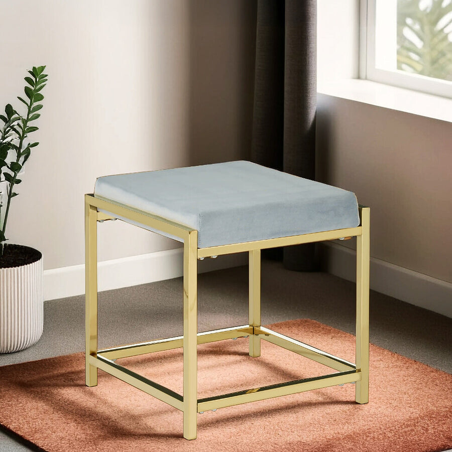 A powder blue velvet stool with a gold metal frame, featuring a square seat and a minimalist design, placed on a textured orange rug in a well-lit room with a window and a potted plant in the background