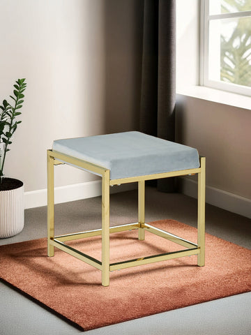 A powder blue velvet stool with a gold metal frame, featuring a square seat and a minimalist design, placed on a textured orange rug in a well-lit room with a window and a potted plant in the background