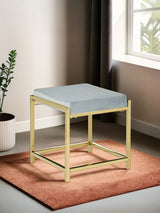 A powder blue velvet stool with a gold metal frame, featuring a square seat and a minimalist design, placed on a textured orange rug in a well-lit room with a window and a potted plant in the background