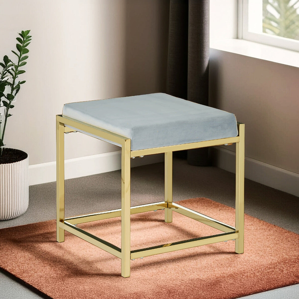 Powder blue velvet stool with a square top and a sleek gold metal frame, positioned on a textured rug in a well-lit room with a window and a potted plant in the background