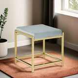 Powder blue velvet stool with a square top and a sleek gold metal frame, positioned on a textured rug in a well-lit room with a window and a potted plant in the background