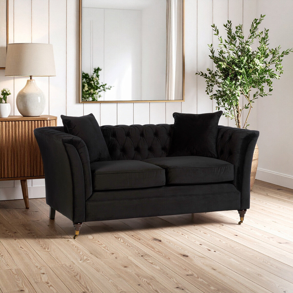 A stylish two-seat sofa in onyx black upholstery featuring tufted backrest and two matching cushions, positioned in a bright living room with a wooden floor, a decorative lamp, a side table, and a potted plant in the background
