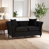 A stylish two-seat sofa in onyx black upholstery featuring tufted backrest and two matching cushions, positioned in a bright living room with a wooden floor, a decorative lamp, a side table, and a potted plant in the background