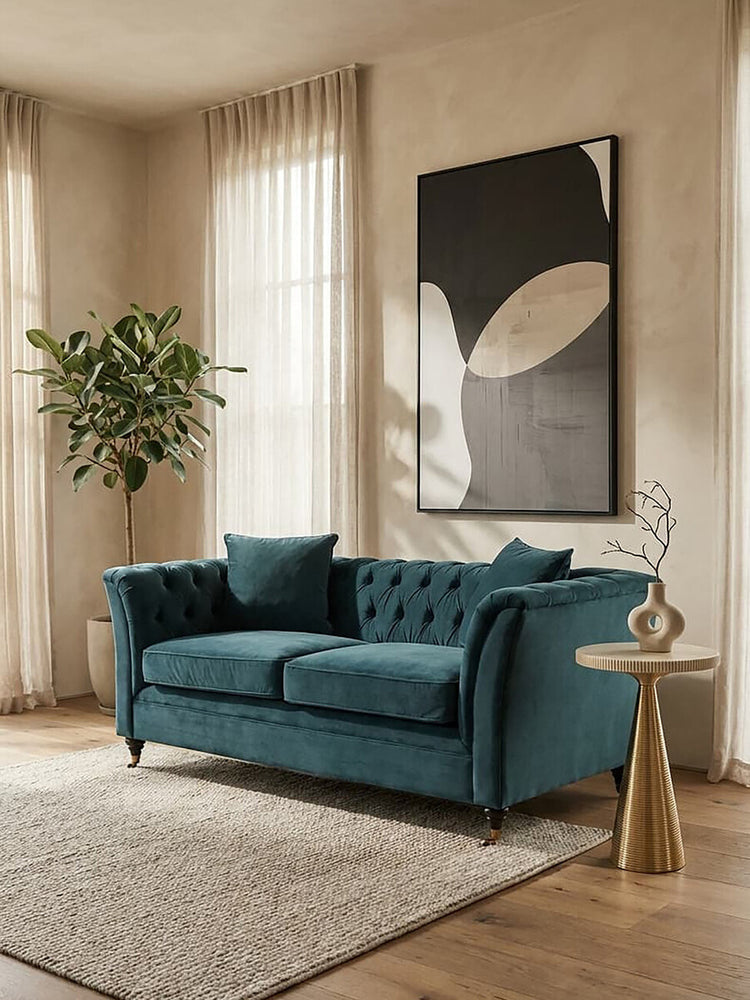 Midnight velvet two-seat sofa with tufted backrest and two matching cushions, placed in a bright room with large windows, a potted plant in the corner, and a modern art piece on the wall. A light-colored area rug is positioned beneath the sofa, and a stylish side table is visible next to it