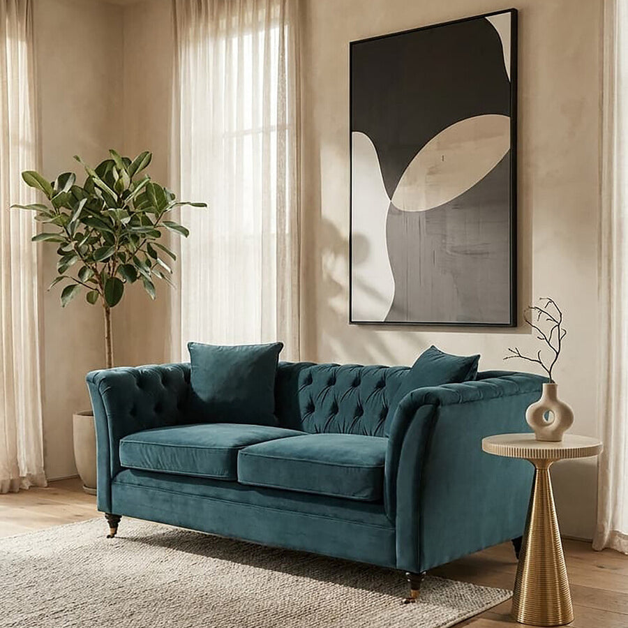 Midnight velvet two-seat sofa with tufted backrest and two matching cushions, placed in a bright room with large windows, a potted plant in the corner, and a modern art piece on the wall. A light-colored area rug is positioned beneath the sofa, and a stylish side table is visible next to it