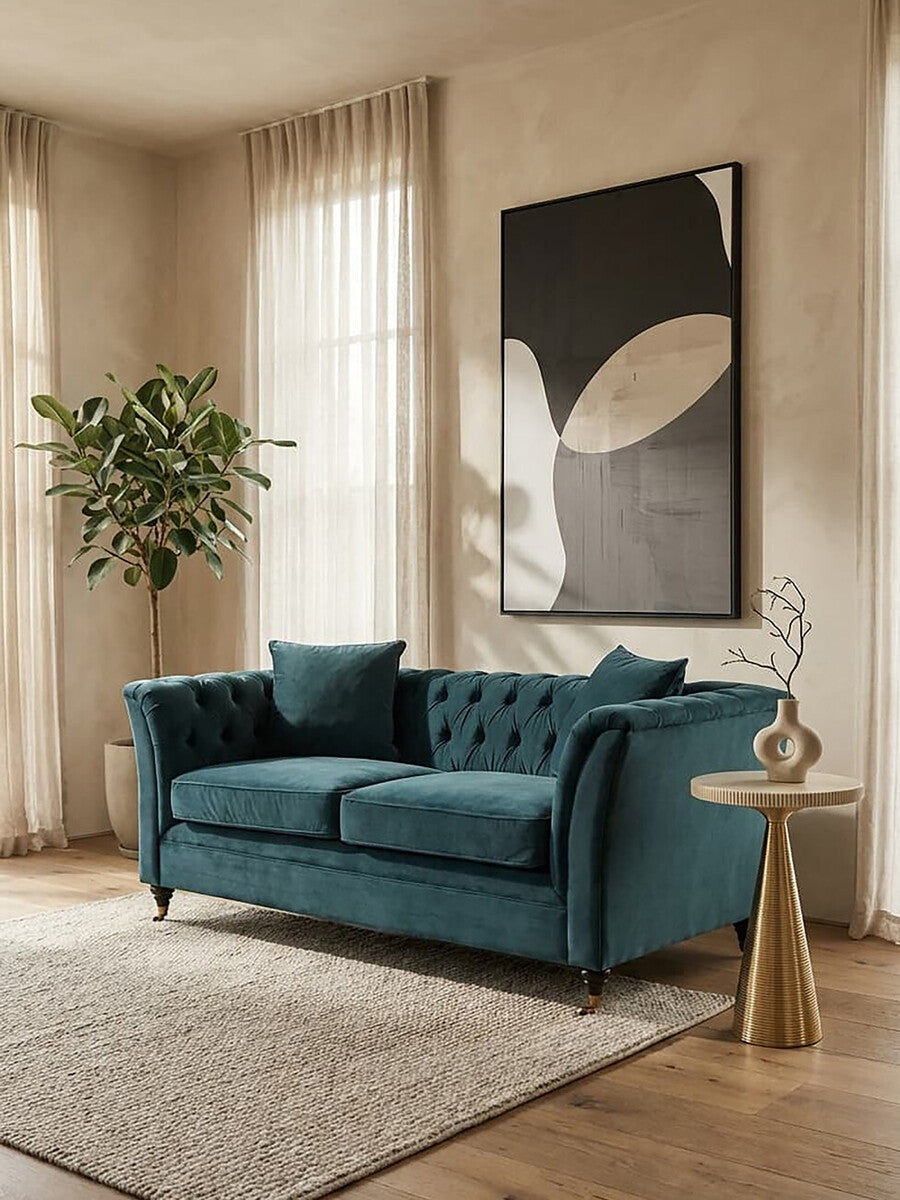 Midnight velvet two-seat sofa with tufted backrest and two matching cushions, placed in a bright room with large windows, a potted plant in the corner, and a modern art piece on the wall. A light-colored area rug is positioned beneath the sofa, and a stylish side table is visible next to it