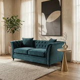 Midnight velvet two-seat sofa with tufted backrest and two matching cushions, positioned in a bright room with large windows, a potted plant in the background, and a small side table next to it. A modern piece of wall art is displayed above the sofa, and a light-colored area rug is on the floor