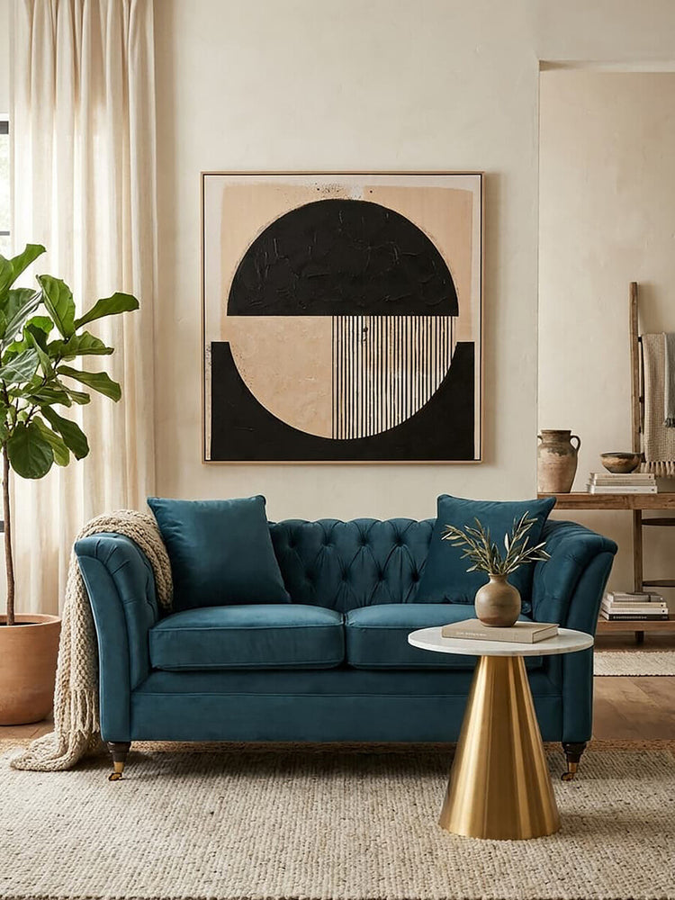 A two-seat sofa upholstered in midnight blue velvet, featuring tufted back cushions and elegant armrests, positioned in a stylish living room setting with a round coffee table and a potted plant. A large abstract wall art piece with geometric shapes in neutral tones is displayed behind the sofa, complemented by soft, light-colored curtains