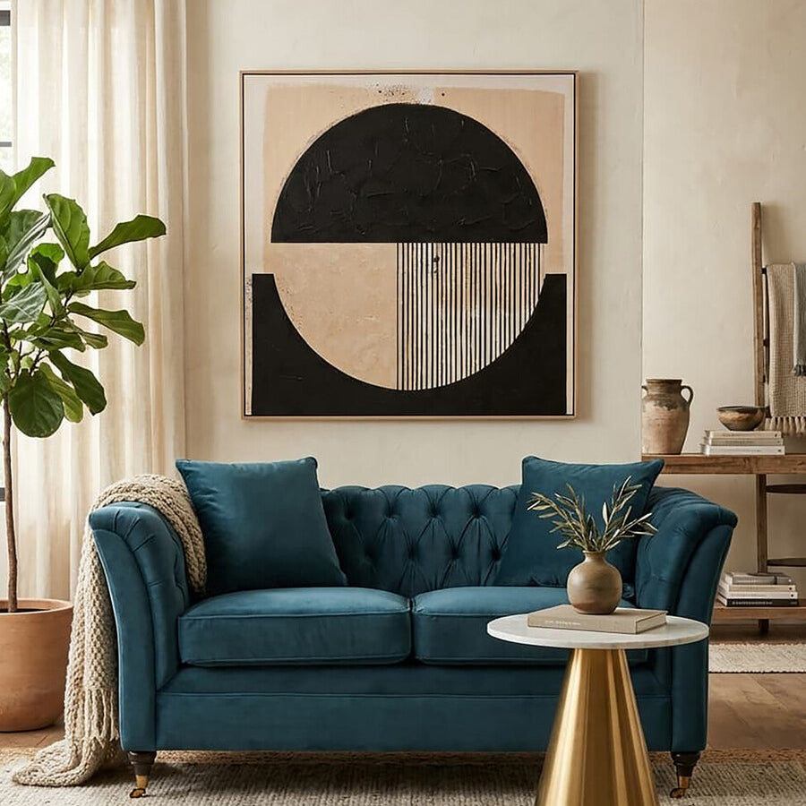 A two-seat sofa upholstered in midnight blue velvet, featuring tufted back cushions and elegant armrests, positioned in a stylish living room setting with a round coffee table and a potted plant. A large abstract wall art piece with geometric shapes in neutral tones is displayed behind the sofa, complemented by soft, light-colored curtains