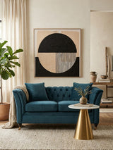A two-seat sofa upholstered in midnight blue velvet, featuring tufted back cushions and elegant armrests, positioned in a stylish living room setting with a round coffee table and a potted plant. A large abstract wall art piece with geometric shapes in neutral tones is displayed behind the sofa, complemented by soft, light-colored curtains