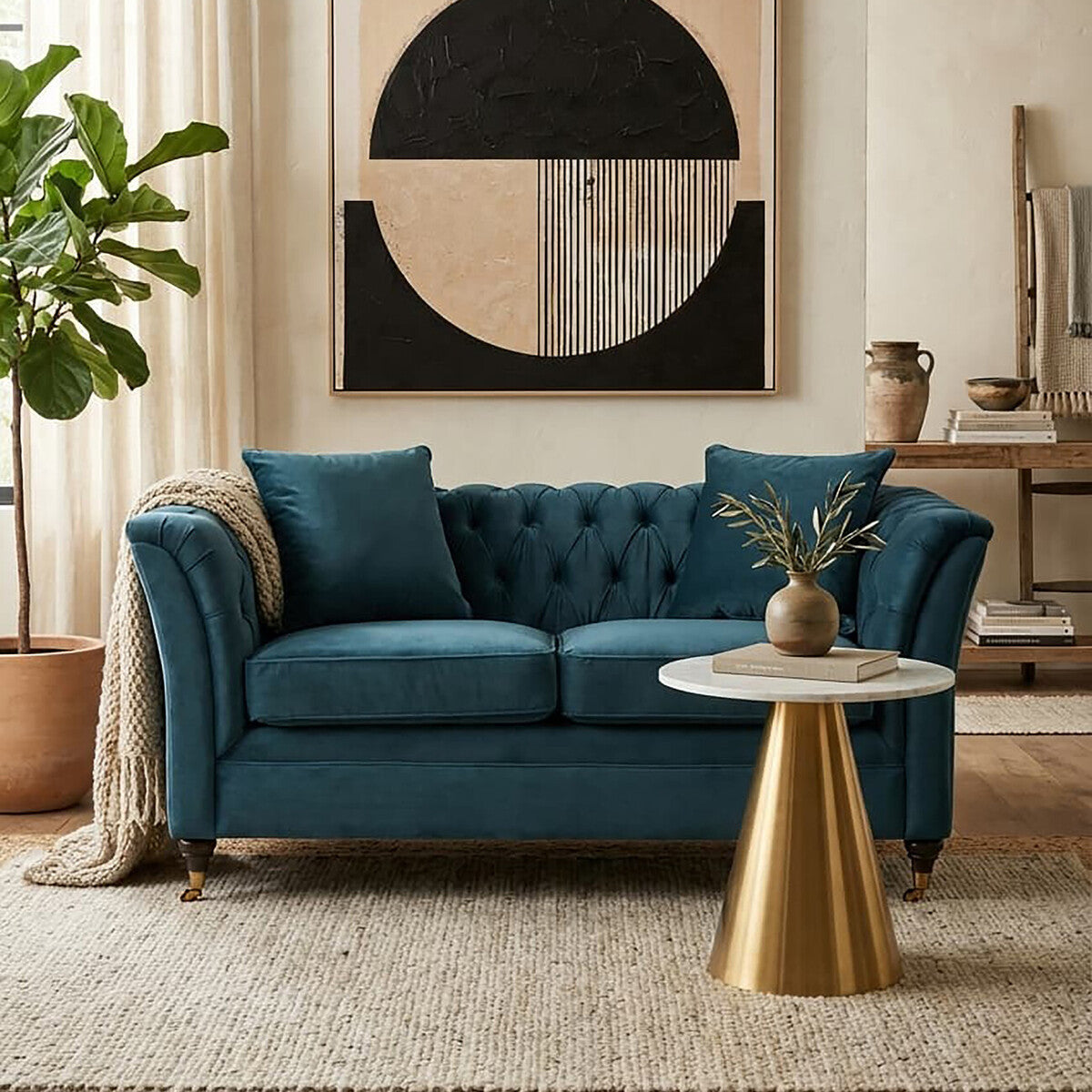 Midnight blue velvet two-seat sofa with tufted backrest and two matching cushions, positioned in a bright room with a large circular wall art piece and a small round gold side table. A potted plant is visible in the background, adding a touch of greenery to the space