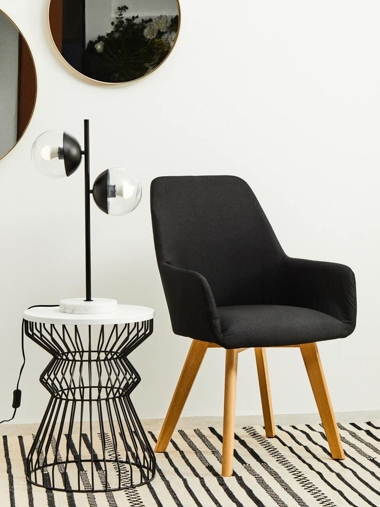 Black fabric accent chair with wooden legs beside wireframe side table and modern table lamp