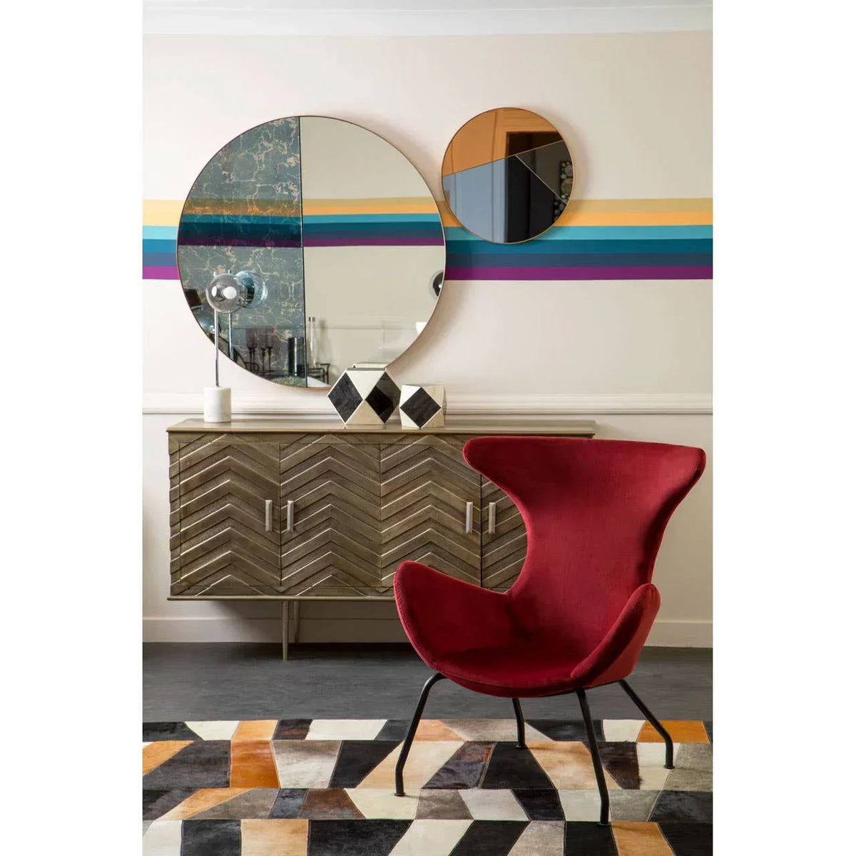 Red accent chair with black legs beside chevron wood sideboard and round wall mirrors