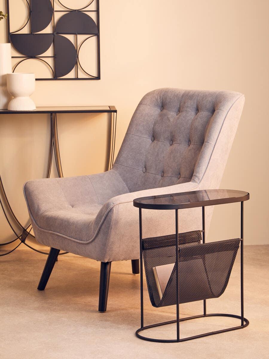 Grey fabric accent chair with button tufting beside black metal side table with magazine rack
