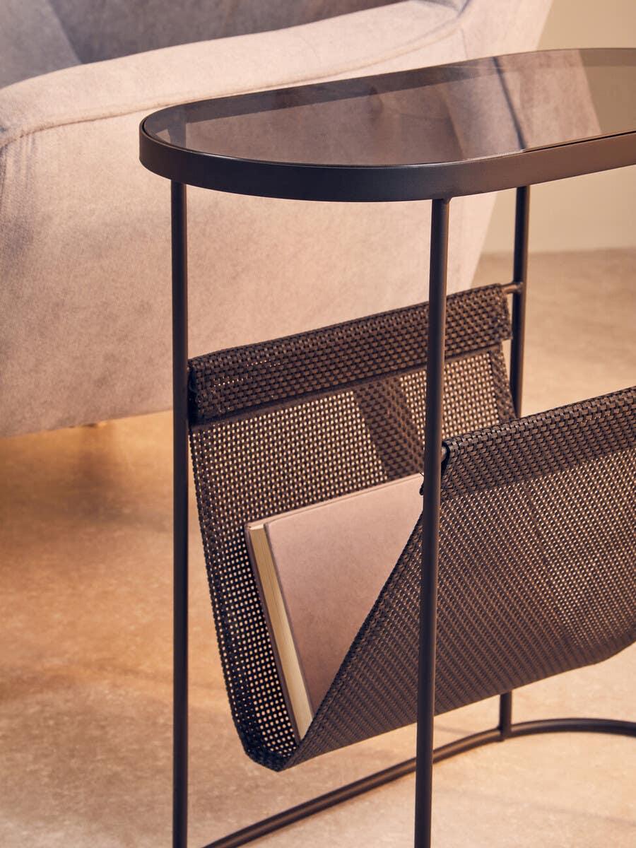 Black metal side table with glass top and mesh magazine holder next to light fabric sofa