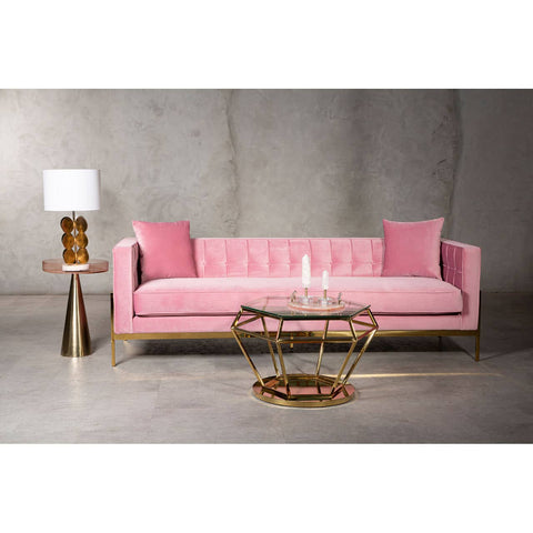 A stylish three-seat sofa upholstered in soft pink fabric, featuring a tufted backrest and two matching cushions. The sofa is supported by a sleek wooden frame with a light finish. In the foreground, a round glass coffee table with a gold metal base is positioned, alongside a modern lamp with a white shade on a side table. The background consists of a textured gray wall, enhancing the contemporary aesthetic of
