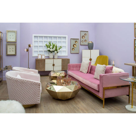 A stylish pink three-seat sofa with wooden legs, placed in a modern living room setting featuring light purple walls, a round gold coffee table, and complementary pastel-colored armchairs. Decorative wall art and a white cabinet with plants add to the cozy atmosphere