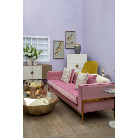 A stylish three-seat pink sofa with wooden legs, adorned with several decorative cushions in various shades of pink and cream, placed against a light purple wall. A round coffee table with a metallic finish sits in front of the sofa, and a vase with fresh flowers is visible on the table. The room features light wooden flooring and framed artwork on the walls