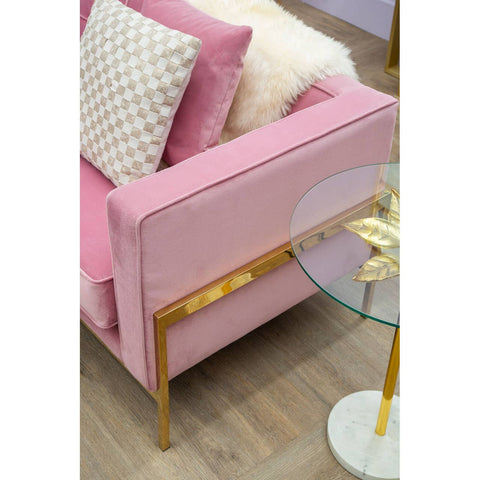 A close-up view of a pink three-seat sofa featuring a sleek design with a gold accent on the armrest. The sofa is adorned with a decorative pillow and is positioned next to a glass side table with a white base. The flooring is wooden, adding warmth to the setting