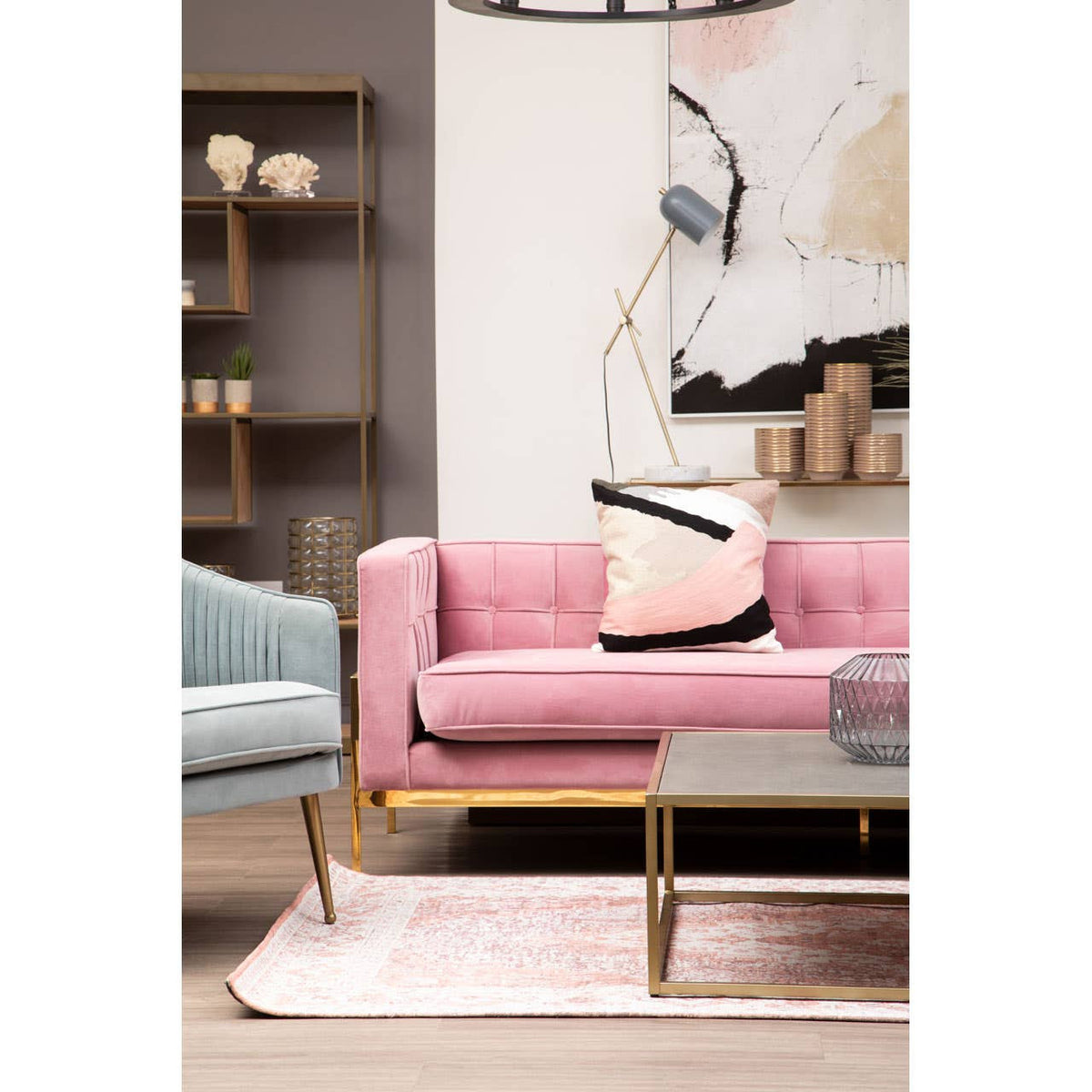 A stylish three-seat sofa in a soft pink fabric, featuring a modern design with gold legs. The sofa is adorned with a decorative black and pink pillow, positioned on a light-colored rug. In the background, there is a minimalist bookshelf and abstract wall art, creating a contemporary living space