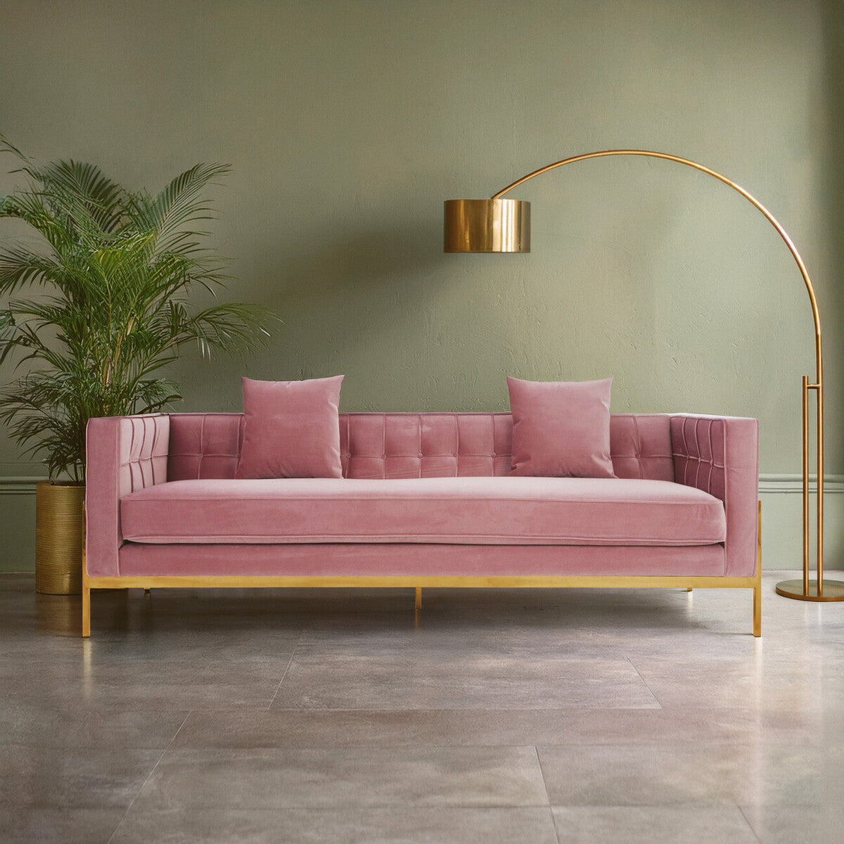 A stylish three-seat pink sofa with a plush fabric finish, featuring two matching cushions, set against a soft green wall. A modern gold arc floor lamp stands beside the sofa, and a leafy potted plant adds a touch of greenery in the background. The floor is made of light-colored tiles, enhancing the contemporary aesthetic