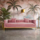 A stylish three-seat pink sofa with a modern design, featuring plush cushions and a sleek wooden frame. The backdrop includes tropical palm trees and a decorative floor lamp, creating a vibrant and inviting living space. The flooring is a polished concrete surface, enhancing the contemporary aesthetic
