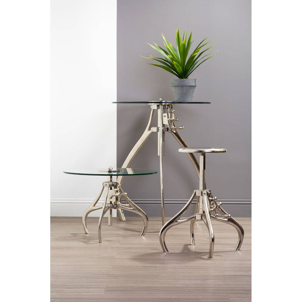 A stylish Maxi Crank Mechanism Stool with a sleek glass top and a unique metal base featuring a modern design, positioned next to a smaller glass table. A potted plant adds a touch of greenery to the scene, set against a neutral wall and wooden floor