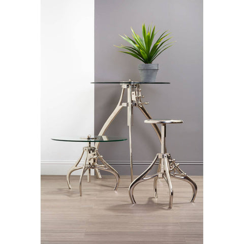A stylish Maxi Crank Mechanism Stool with a sleek glass top and a unique metal base featuring a modern design, positioned next to a smaller glass table. A potted plant adds a touch of greenery to the scene, set against a neutral wall and wooden floor