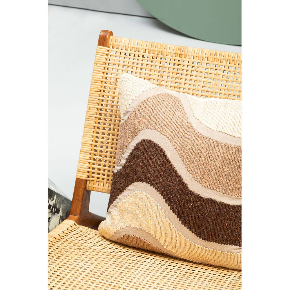 A close-up view of the Lovina Wood and Natural Rattan Lounge Chair featuring a woven rattan backrest and seat. A decorative pillow with a wave pattern in shades of beige, brown, and cream is placed on the chair, adding a cozy touch to the natural materials. The background includes a hint of greenery, enhancing the relaxed atmosphere